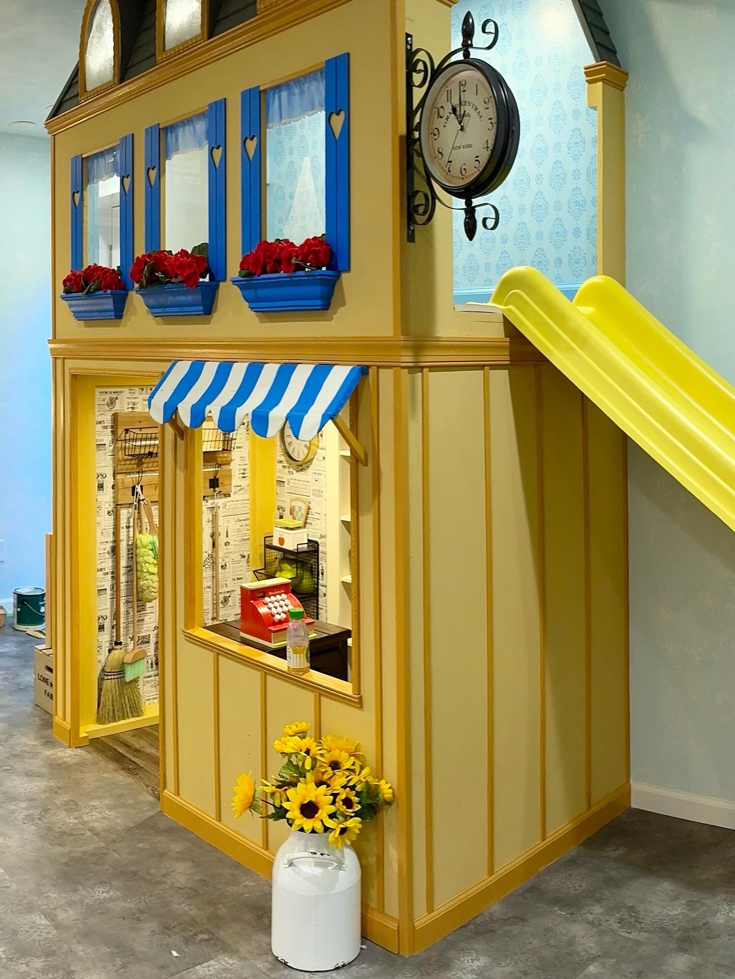 Flower shop playhouse: clock tower, blue shutters, striped awning, and newspaper wallpaper interior