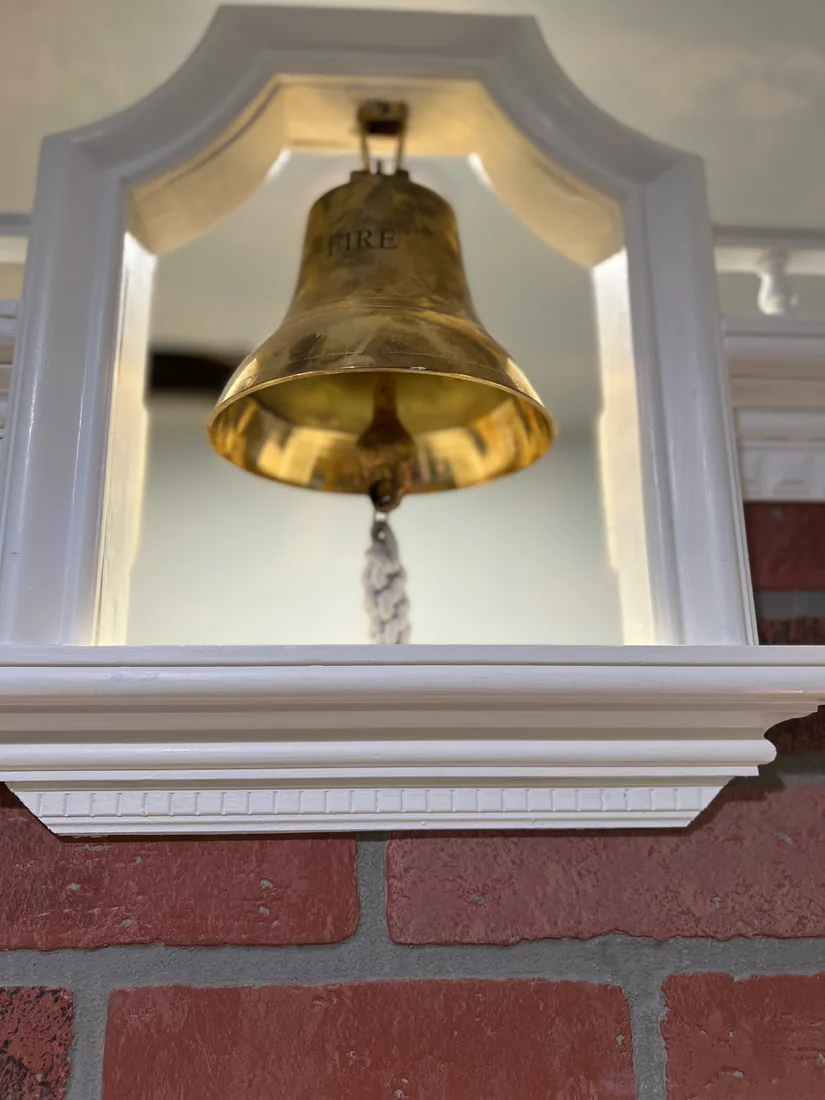 A polished brass bell hanging in a white pediment housing over the Fire Station's brick facade